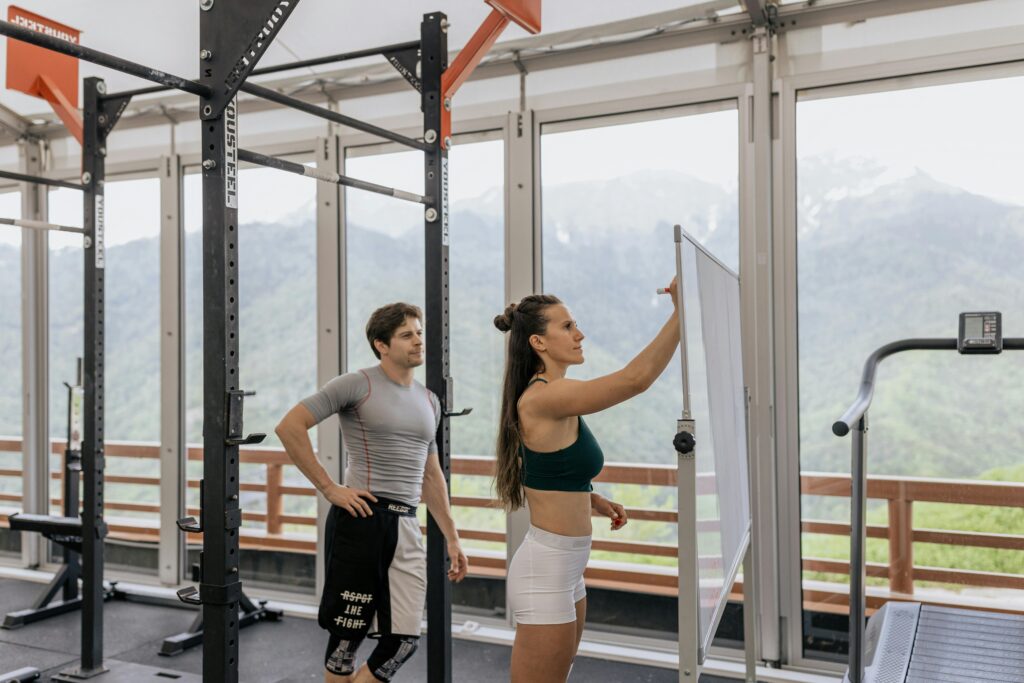 A gym coach instructs on whiteboard beside fitness equipment with mountain view.